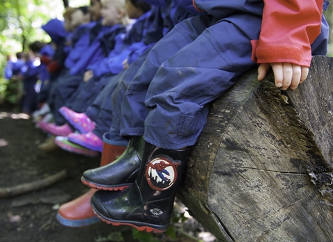 Children in matching rain suits sitting in a row on a log in the middle of a woodland. Just their legs are in shot