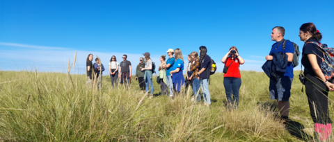 A group of people standing in a line across a grassy sand dune and looking into the distance. The sky is bright blue