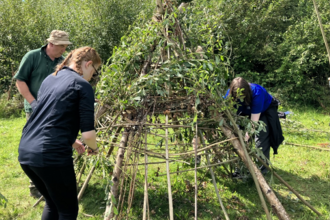 Two young people weave willow around stakes in the ground to make a stone age style teepee house