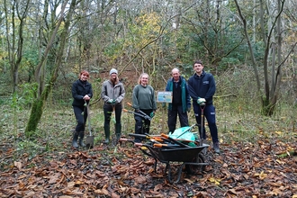 A group of 5 volunteers stood in a woodland next to a wheelbarrow of tools