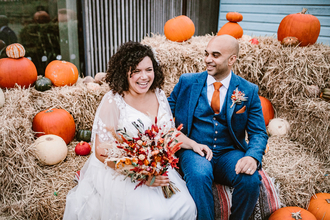 A bride and groom sit on hay bales surrounded by pumpkins