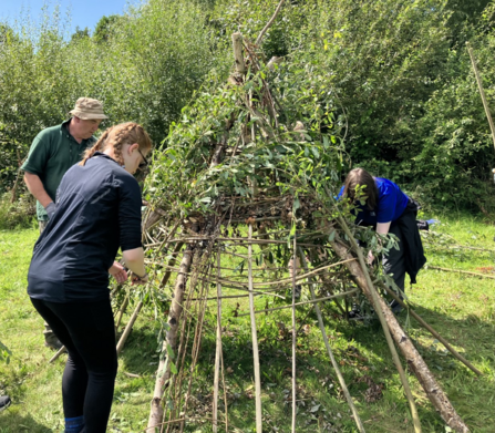 Two young people weave willow around stakes in the ground to make a stone age style teepee house