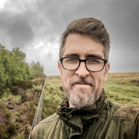 A man wearing glasses stands on a moor looking at the camera