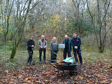 A group of 5 volunteers stood in a woodland next to a wheelbarrow of tools