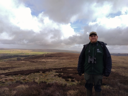 A man wearing a green fleece with binoculars standing on a moor