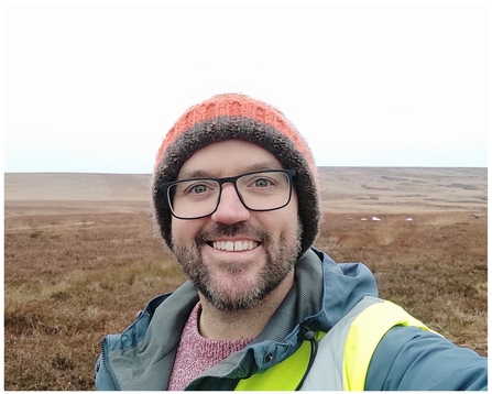 A man wearing a wooly hat and glasses with a short beard standing on a moor